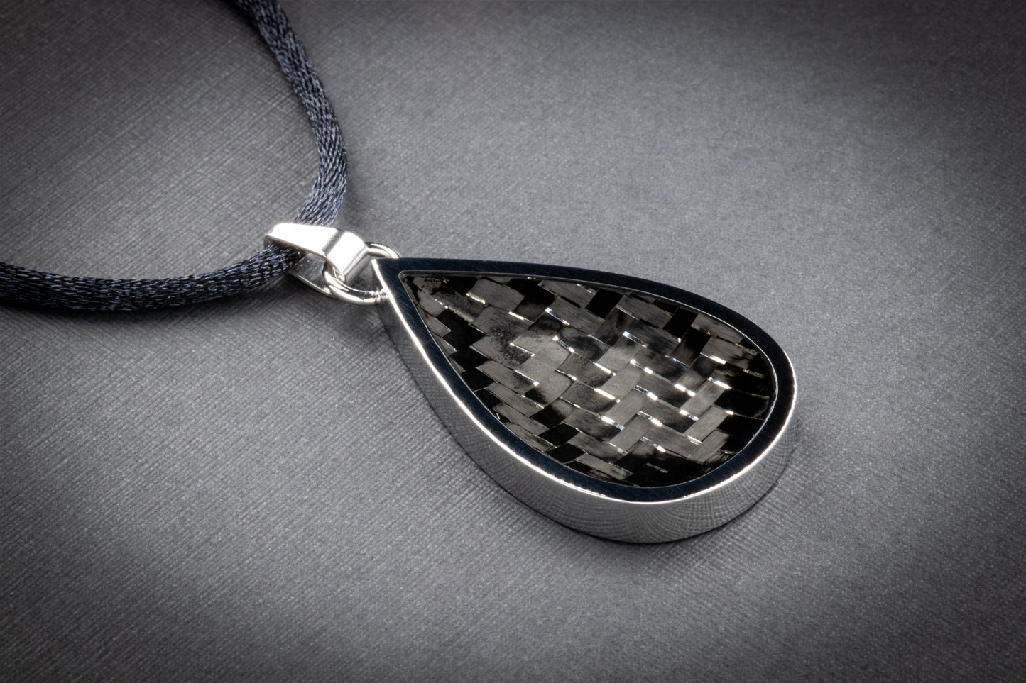 Tropfenförmiger Anhänger mit einer polierten Edelstahlfassung und einer Einlage aus klassisch gewebtem Carbon in Schwarz-Grau. Die Oberfläche reflektiert das Licht leicht und zeigt die charakteristische Struktur des Carbonmaterials – technisch, elegant und zeitlos. Der Anhänger hängt an einem dunkelgrauen, textilummantelten Band und liegt auf einer dezent strukturierten grauen Unterlage. Ein puristisches, hochwertiges Schmuckstück von Regenstein.shop – für alle, die minimalistisches Design mit Hightech-Mate