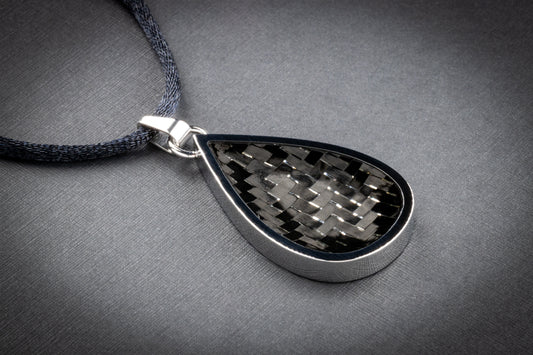 Tropfenförmiger Anhänger mit einer polierten Edelstahlfassung und einer Einlage aus klassisch gewebtem Carbon in Schwarz-Grau. Die Oberfläche reflektiert das Licht leicht und zeigt die charakteristische Struktur des Carbonmaterials – technisch, elegant und zeitlos. Der Anhänger hängt an einem dunkelgrauen, textilummantelten Band und liegt auf einer dezent strukturierten grauen Unterlage. Ein puristisches, hochwertiges Schmuckstück von Regenstein.shop – für alle, die minimalistisches Design mit Hightech-Mate