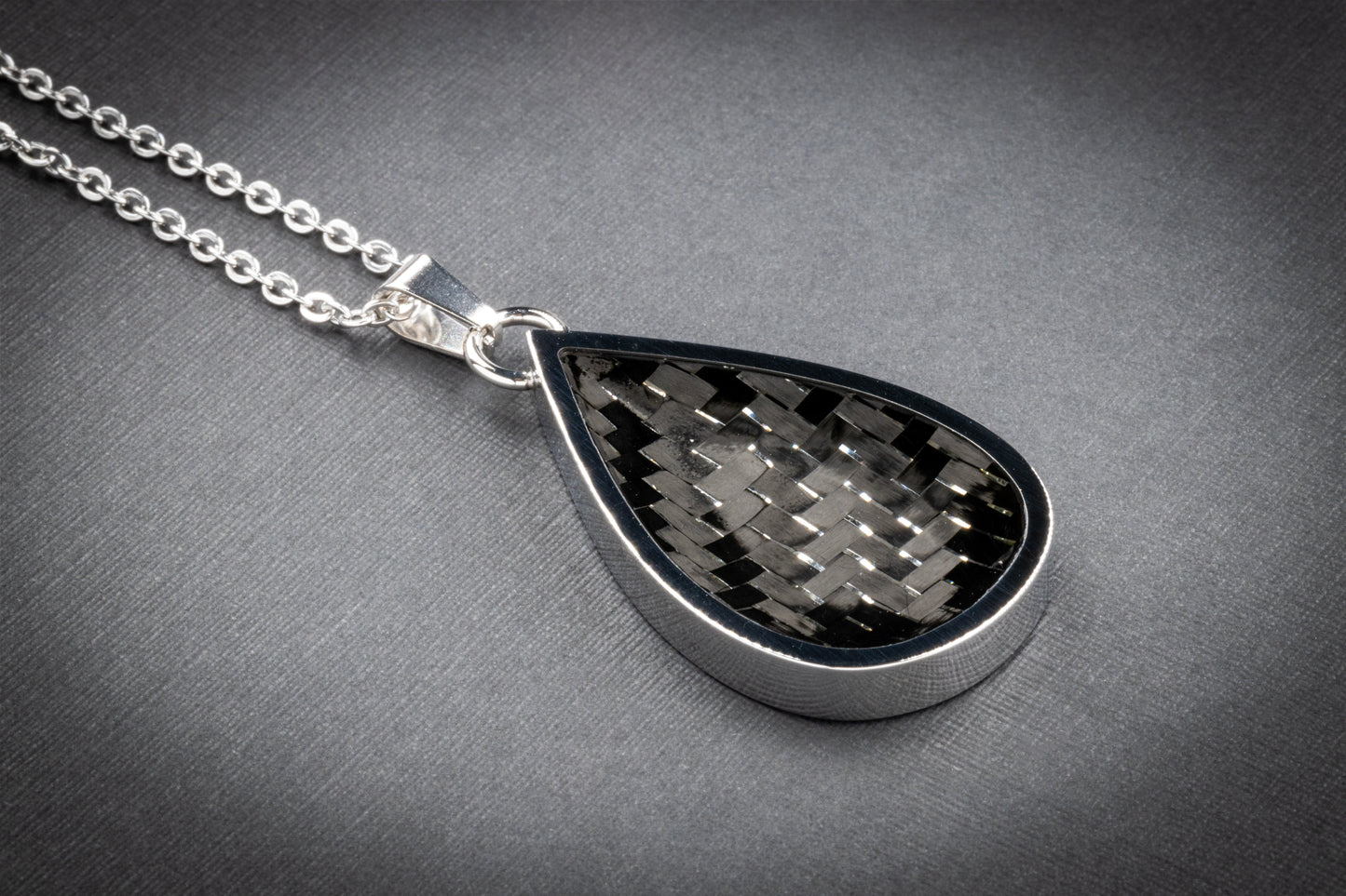 Tropfenförmiger Anhänger mit glänzender Edelstahlfassung und einer Einlage aus klassisch gewebtem Carbon in Schwarz-Grau, ganz ohne farbige Akzente. Der technische Look wirkt minimalistisch und zeitlos zugleich. Der Anhänger ist an einer silberfarbenen Gliederkette befestigt und liegt auf einer fein strukturierten, grauen Oberfläche. Ideal für Liebhaber:innen schlichter Eleganz – ein hochwertiges Designstück von Regenstein.shop.