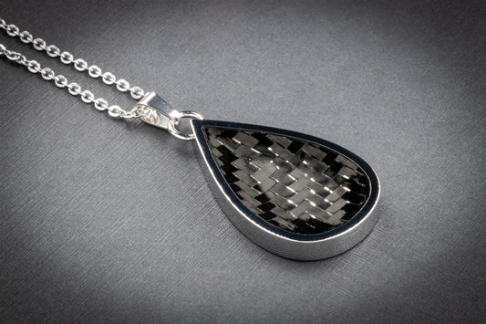 Tropfenförmiger Anhänger mit glänzender Edelstahlfassung und einer Einlage aus klassisch gewebtem Carbon in Schwarz-Grau, ganz ohne farbige Akzente. Der technische Look wirkt minimalistisch und zeitlos zugleich. Der Anhänger ist an einer silberfarbenen Gliederkette befestigt und liegt auf einer fein strukturierten, grauen Oberfläche. Ideal für Liebhaber:innen schlichter Eleganz – ein hochwertiges Designstück von Regenstein.shop.