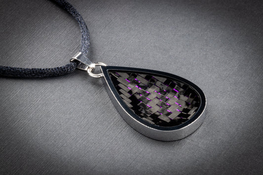 Tropfenförmiger Anhänger mit einer eleganten Fassung aus poliertem Edelstahl. Die Einlage besteht aus gewebtem Carbon mit feinen, violett schimmernden Fasern, die im Licht dezent reflektieren und dem Schmuckstück eine moderne, futuristische Ausstrahlung verleihen. Der Anhänger ist an einem dunkelgrauen, textilummantelten Band befestigt und liegt auf einer strukturierten grauen Oberfläche. Ein stilvolles Designerstück von Regenstein.shop – bekannt für hochwertigen Schmuck mit innovativen Materialien.