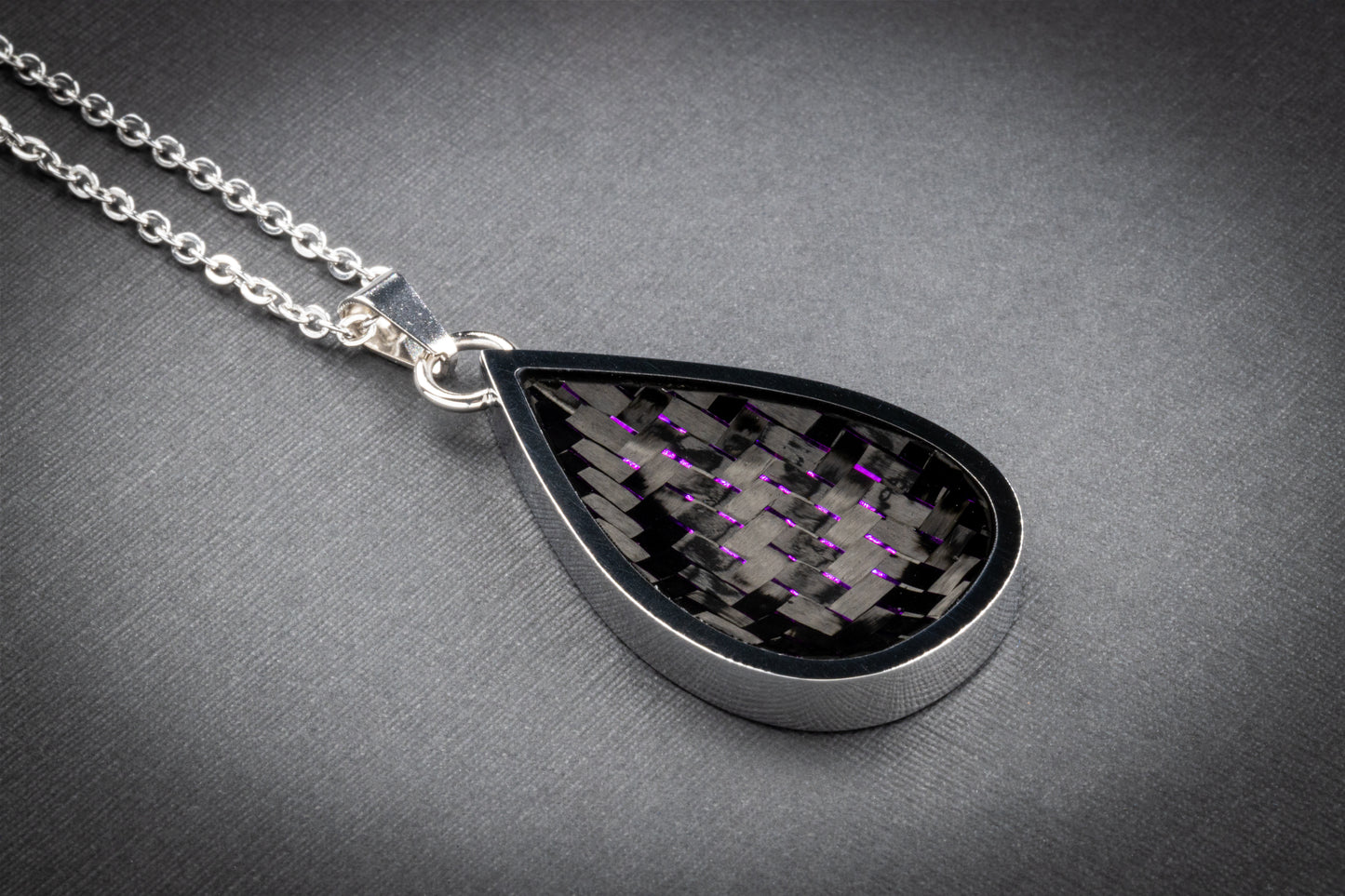 Tropfenförmiger Anhänger aus glänzendem Edelstahl mit einer Einlage aus gewebtem Carbon, durchzogen von fein schimmernden violetten Fasern. Das auffällige Farbspiel verleiht dem technisch inspirierten Material eine elegante, leicht futuristische Note. Der Anhänger ist an einer filigranen, silberfarbenen Gliederkette befestigt und liegt auf einer dezent strukturierten, grauen Oberfläche. Ein einzigartiges Schmuckstück aus der Kollektion von Regenstein.shop – hochwertiger Schmuck für Individualist:innen.