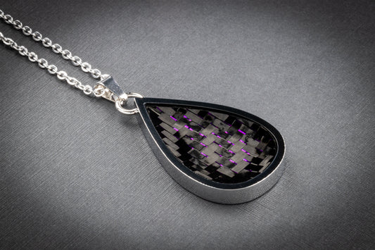 Tropfenförmiger Anhänger aus glänzendem Edelstahl mit einer Einlage aus gewebtem Carbon, durchzogen von fein schimmernden violetten Fasern. Das auffällige Farbspiel verleiht dem technisch inspirierten Material eine elegante, leicht futuristische Note. Der Anhänger ist an einer filigranen, silberfarbenen Gliederkette befestigt und liegt auf einer dezent strukturierten, grauen Oberfläche. Ein einzigartiges Schmuckstück aus der Kollektion von Regenstein.shop – hochwertiger Schmuck für Individualist:innen.